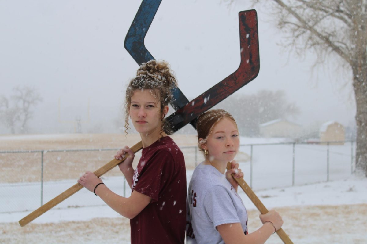 Photo of Kyra Koppleman and Lula Hobbs holding up hockey sticks in the snow.